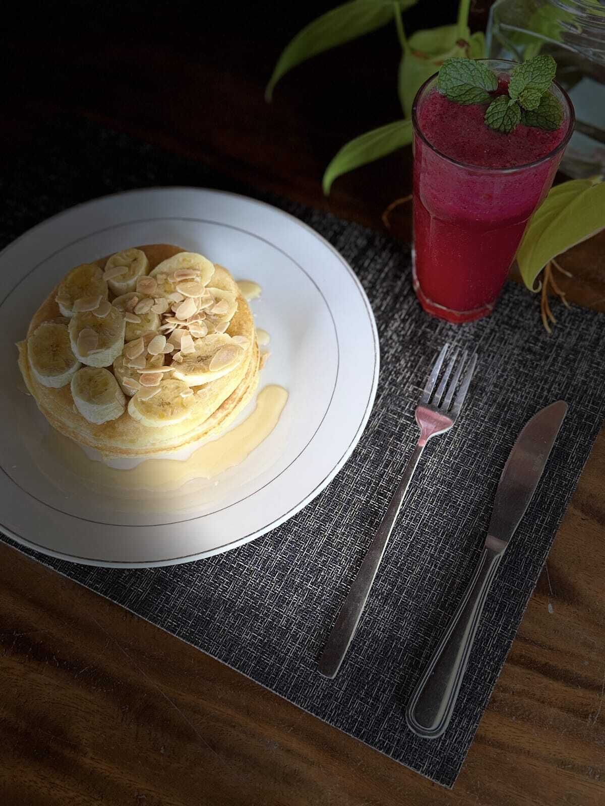 A plate of pancakes topped with sliced bananas and almonds, a glass of red beverage with a mint garnish, and cutlery on a textured placemat.