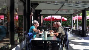 An outdoor cafe with tables and chairs, red umbrellas, and potted plants. - Peaceful Apartment With Pool (Turnhout)