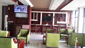 Un vestíbulo de hotel con sillones tapizados de verde, un televisor mostrando noticias y una zona de bar con un reloj y detalles decorativos.