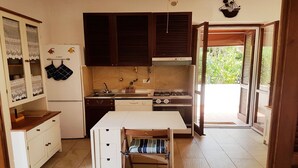 Eine kompakte Küche mit Herd, Spüle und Schränken. Ein Fenster mit Jalousien, ein Kühlschrank und ein kleiner Esstisch mit Stuhl sind ebenfalls vorhanden.