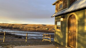 A rustic cabin with a wooden door and window, a wooden deck, and a view of a river and hills in the background. - Classy Hillside Tiny House (Monarch)
