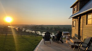 Una terraza de madera con sillas y una parrilla, con vista a un río y campos durante el atardecer.