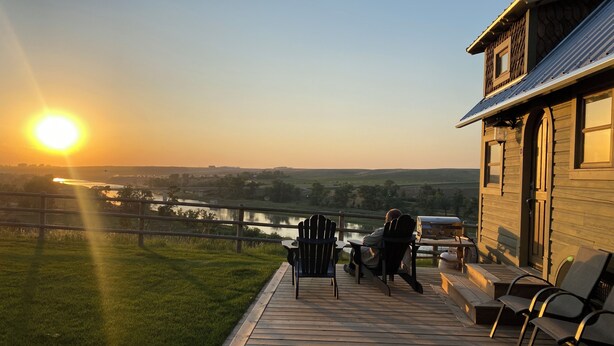 A wooden deck with chairs and a grill, overlooking a river and fields during sunset. - Classy Hillside Tiny House (Monarch)