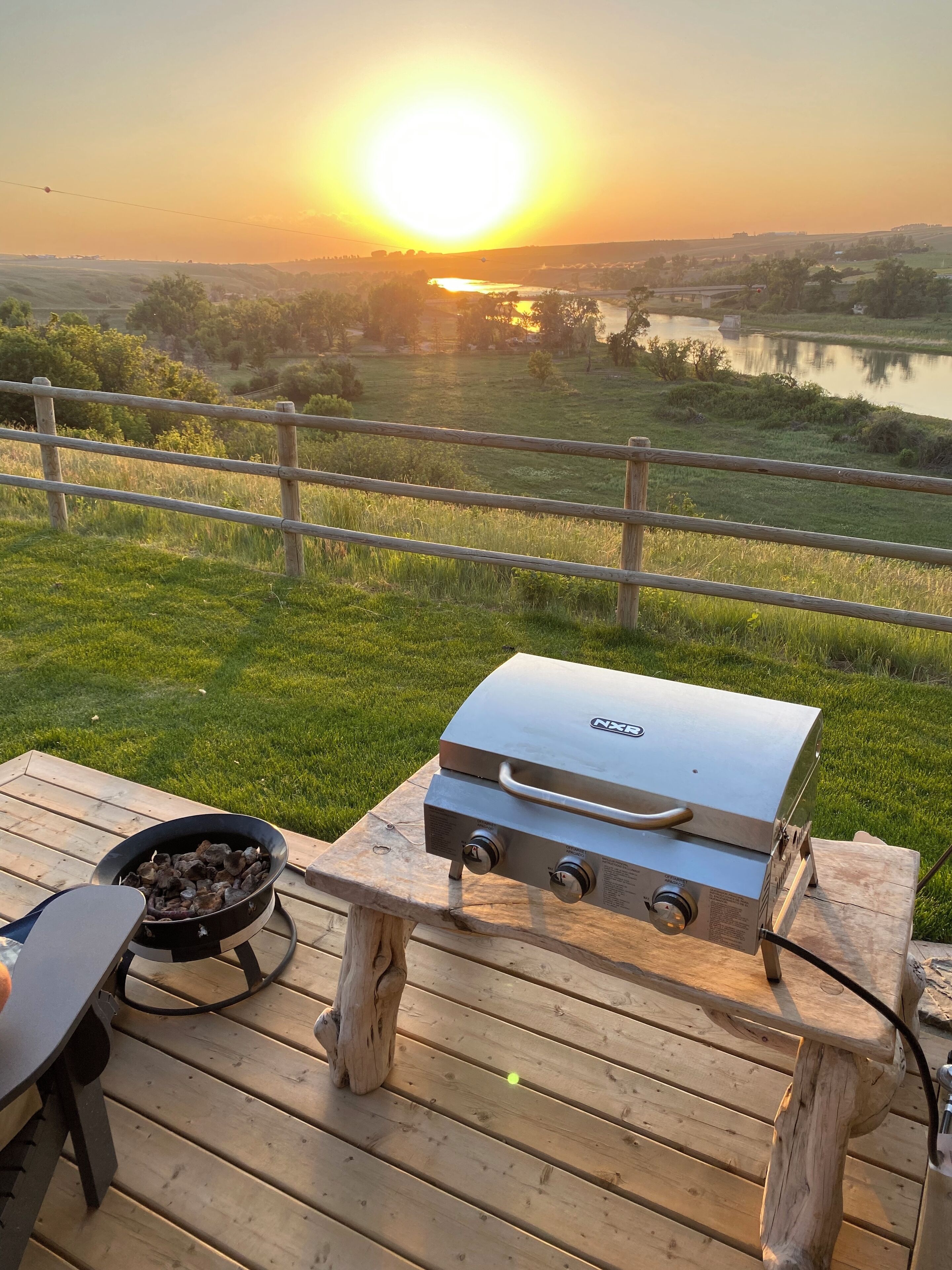 A wooden deck with a grill and a pan, overlooking a scenic landscape with a sunset.