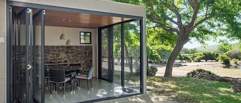 A modern outdoor dining area with a stone wall, glass doors, and a view of a tree-lined landscape.