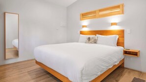 A modern bedroom with a wooden bed frame, white bedding, a bedside table, and a mirror.