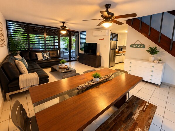 A modern living room with a wooden dining table, a black sofa, and a kitchen area with a refrigerator and a TV.