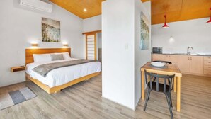 A bedroom with a wooden bed, a bedside table, a nightstand, a window with a view, and a kitchen area with a sink and cabinets.