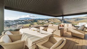 A covered outdoor seating area with wicker furniture and a table, overlooking a scenic landscape with hills and buildings.