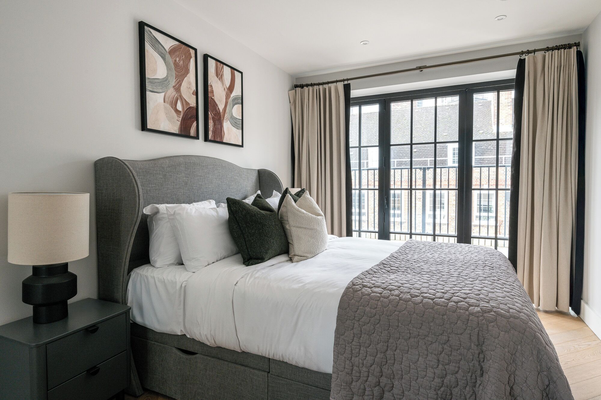 Una camera da letto con un'ampia finestra, un letto con biancheria bianca, un poggiatesta grigio e due opere d'arte incorniciate appese al muro.