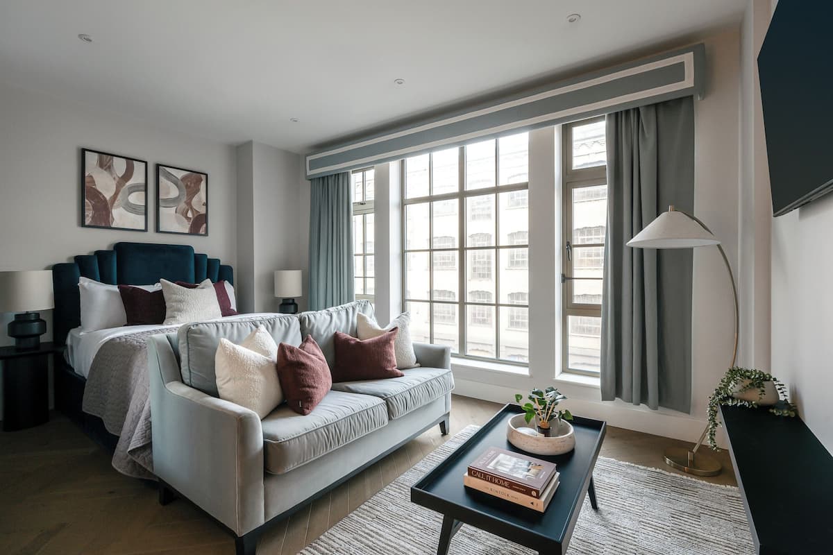 Habitación de hotel moderna con una cama grande, un sofá gris, una mesa de centro con libros y vistas amplias a través de grandes ventanales.