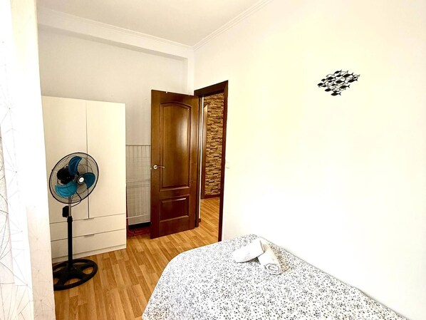 A bedroom with a bed, a standing fan, a wardrobe, and a door leading to another room.