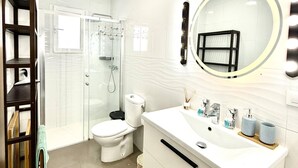 Um banheiro moderno com chuveiro em cabine de vidro, vaso sanitário branco, toucador com espelho grande e pia com artigos de higiene pessoal.