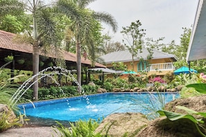 A swimming pool with a water feature, surrounded by tropical plants and a building with a tiled roof.