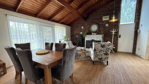 A dining area with a wooden table and chairs, a brick fireplace, and a large window with curtains. - 3 Bdrm Farm Stay with Goats, Hairy Coos and chickens. Farm Life. You'll Love it! (Postmans Ridge)