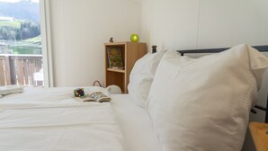 A neatly made bed with white linens, a bedside table with a book and a Rubik's cube, a wooden nightstand with a lamp and a book, and a window with a view of greenery.