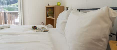 A neatly made bed with white linens, a small bedside table with a book and a Rubik's cube, and a wooden nightstand with a lamp and a book.