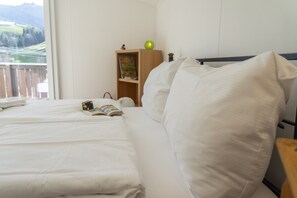 Un lit bien fait, avec du linge de lit blanc, une table de chevet sur laquelle se trouvent un livre et un Rubik's Cube, une commode en bois avec une lampe et un livre, et une fenêtre donnant sur un paysage verdoyant.