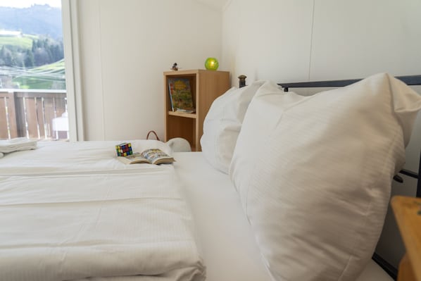 A neatly made bed with white linens, a small bedside table with a book and a Rubik's cube, and a wooden nightstand with a lamp and a book. - Füchsle holiday flat with mountain views in Wildhaus (Wildhaus)