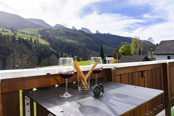 A table with two wine glasses and a glass of water, a pair of sunglasses, and a wooden railing overlooking a mountainous landscape.