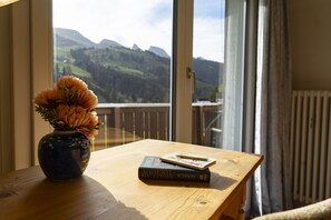 A wooden table with a vase of flowers and a book, near a window with a view of mountains.