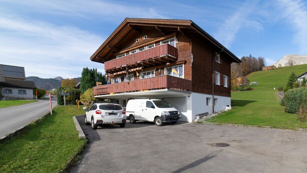 A two-story building with a balcony, a parking area with two cars, and a clear sky.