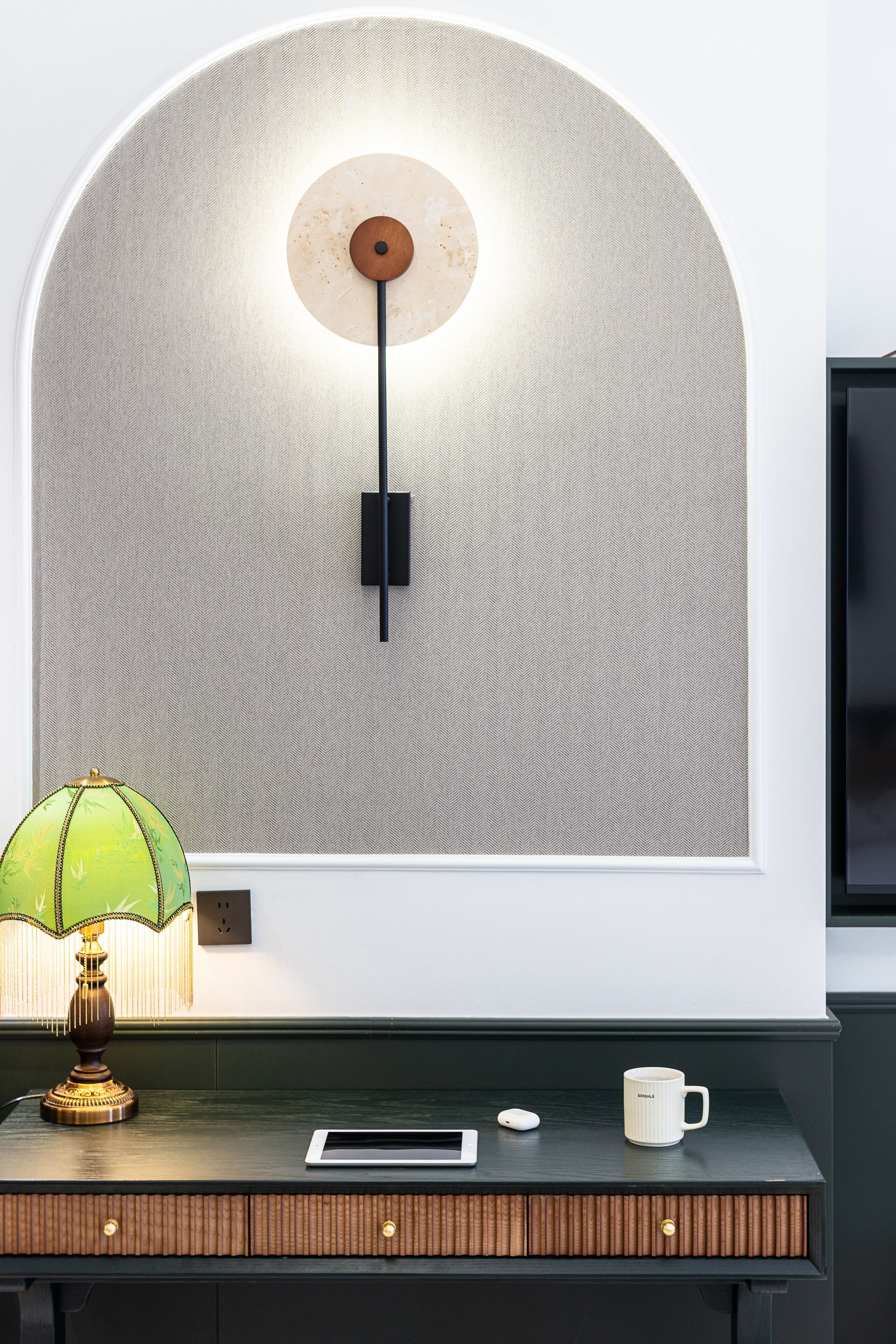 A modern room with a desk, a lamp, a wall-mounted light fixture, and a flat-screen TV.