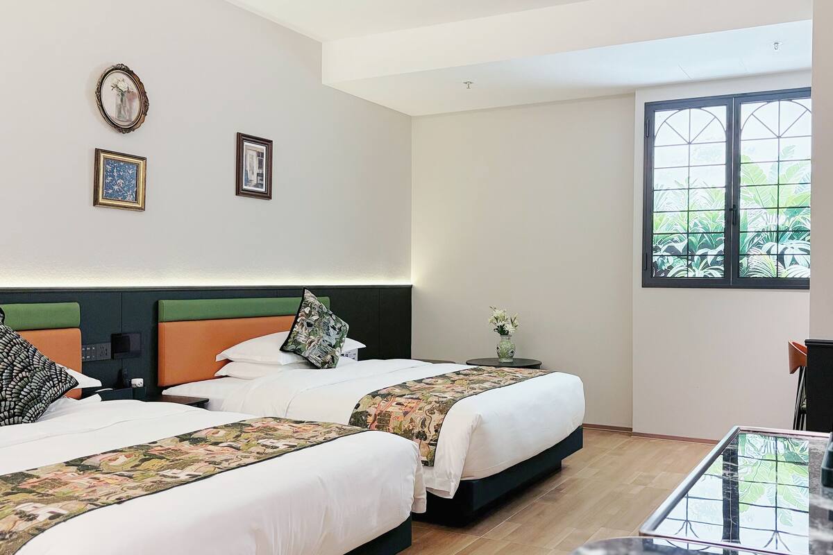 Una habitación de hotel con dos camas, una mesita con una cafetera, un ventanal con vistas a la vegetación y una pared con dos cuadros enmarcados.