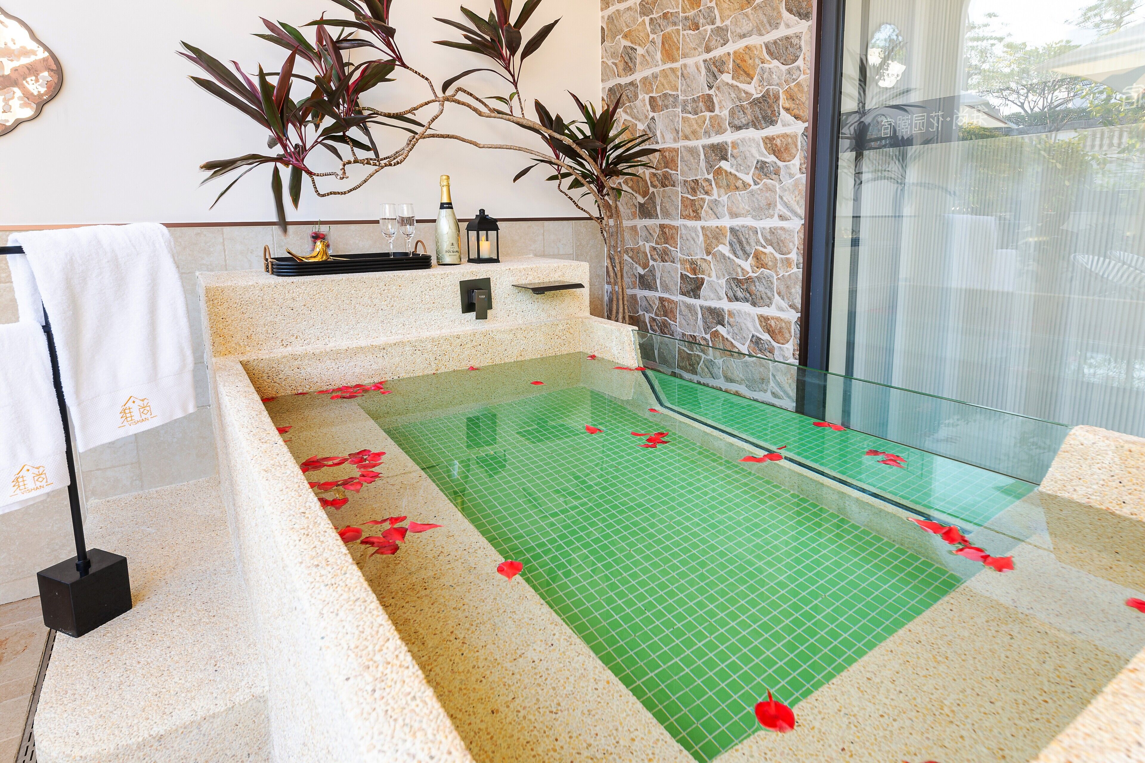 A spa area with a stone wall, a glass-enclosed hot tub, and a small shelf with bottles and glasses.