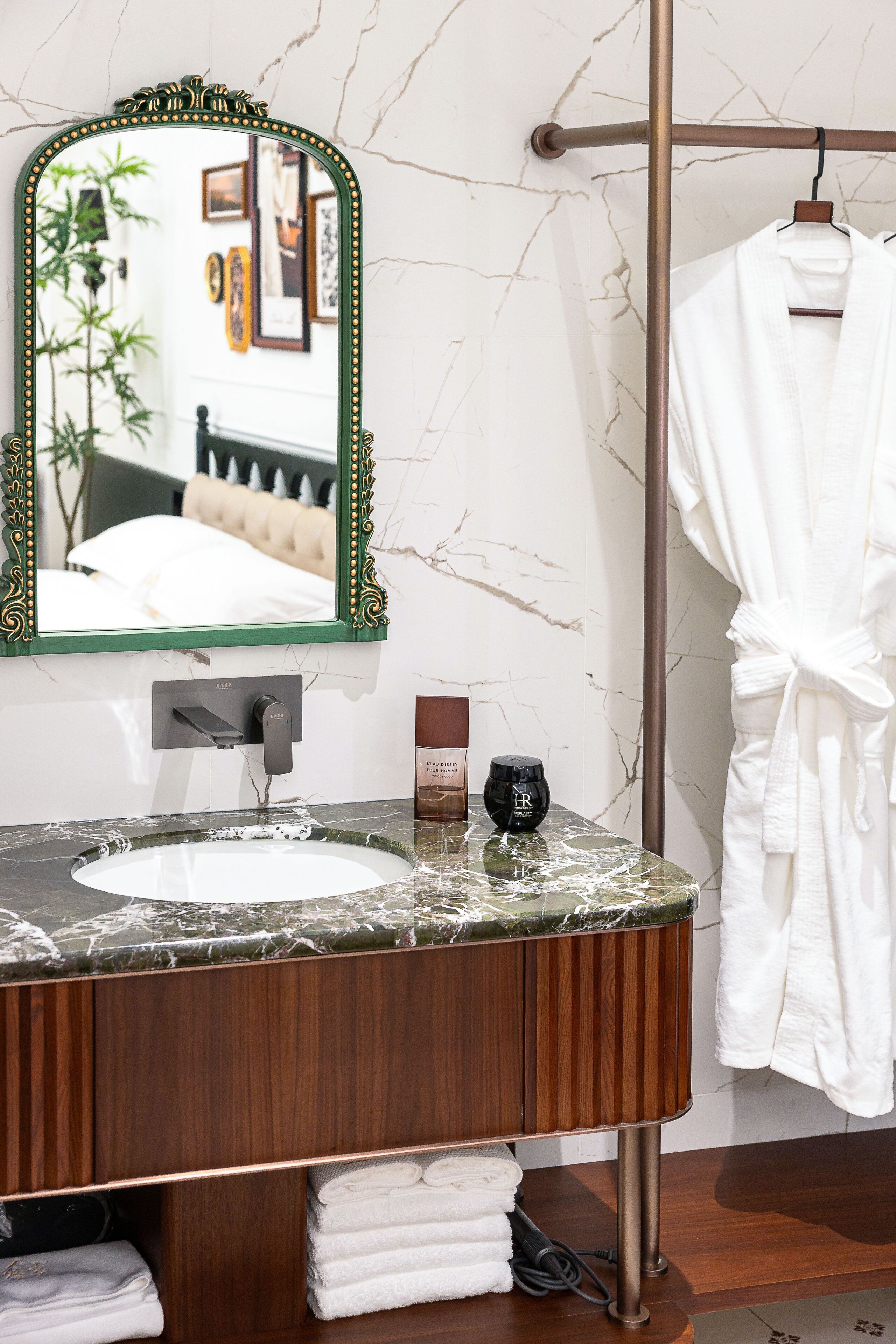 A bathroom with a marble sink, a mirror with a green frame, a towel rack with a robe, and a wooden vanity.