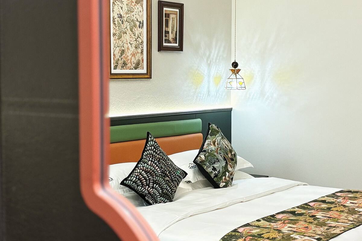 Una cama bien hecha con una colcha estampada, dos almohadas decorativas y un cabecero tapizado en verde.