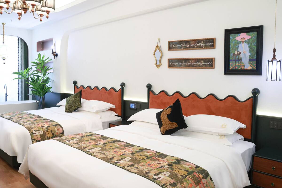 Habitación de hotel con dos camas, ropa de cama blanca, almohadones decorativos, una planta grande y obras de arte en la pared.