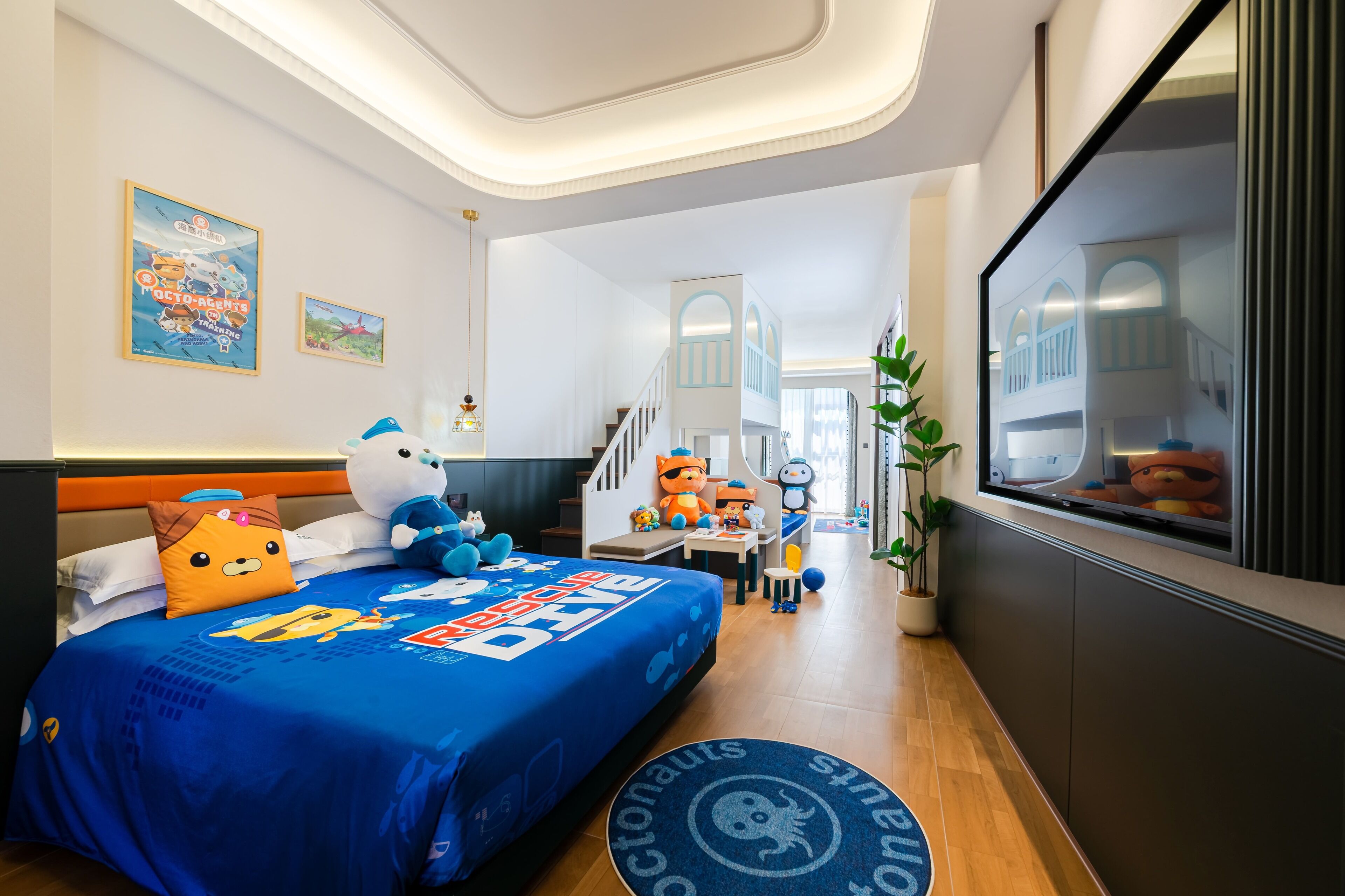 Una habitación de hotel con una cama cubierta con una colcha azul adornada con personajes de dibujos animados, un ventanal con vistas a un balcón y una planta en maceta junto a la ventana.