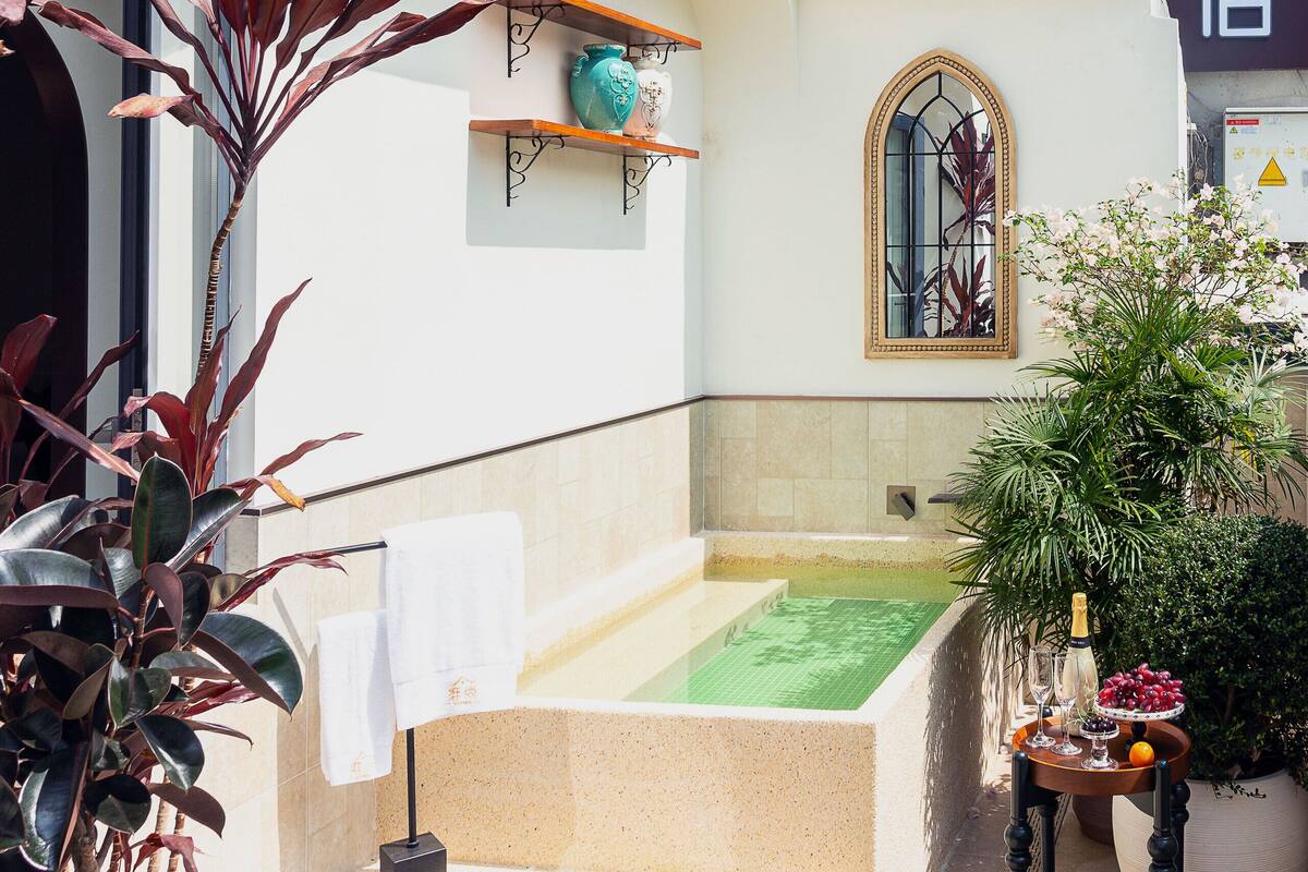 Entrada de hotel con piscina, plantas en macetas y un cartel en idioma extranjero.