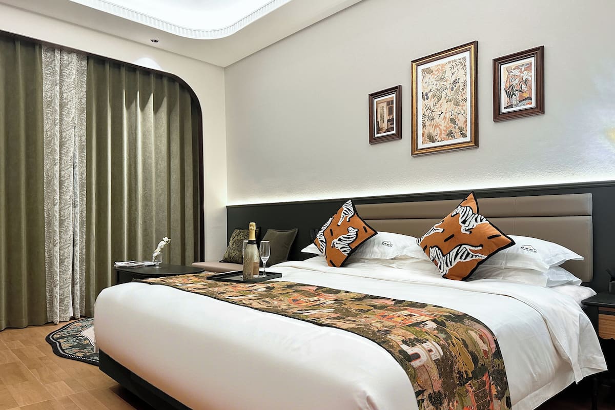 Habitación de hotel con una cama grande, una mesita de noche, un edredón decorativo, un cuadro enmarcado y una cortina.