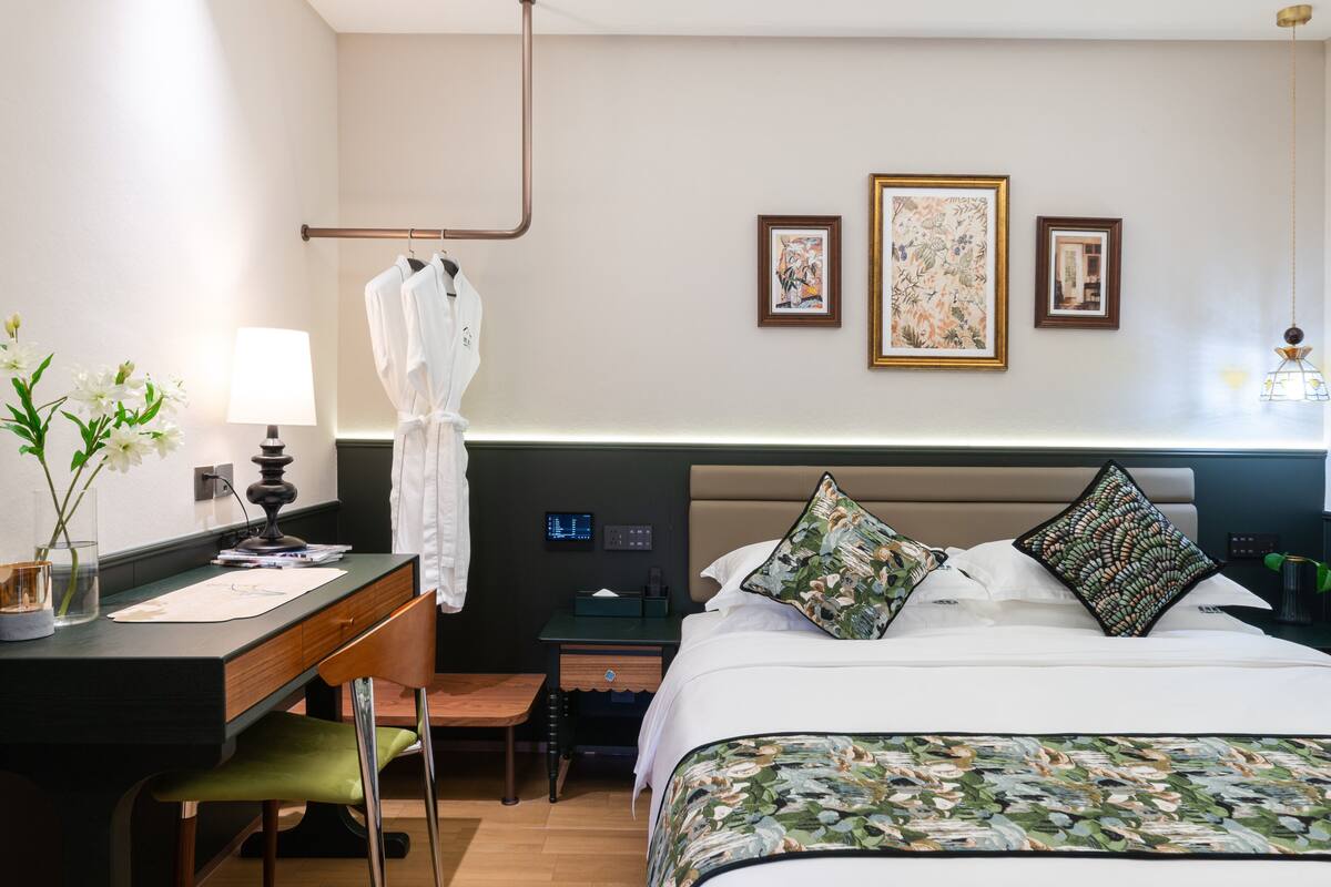 Una habitación de hotel con cama, escritorio, silla, perchero para bata y obras de arte en la pared.