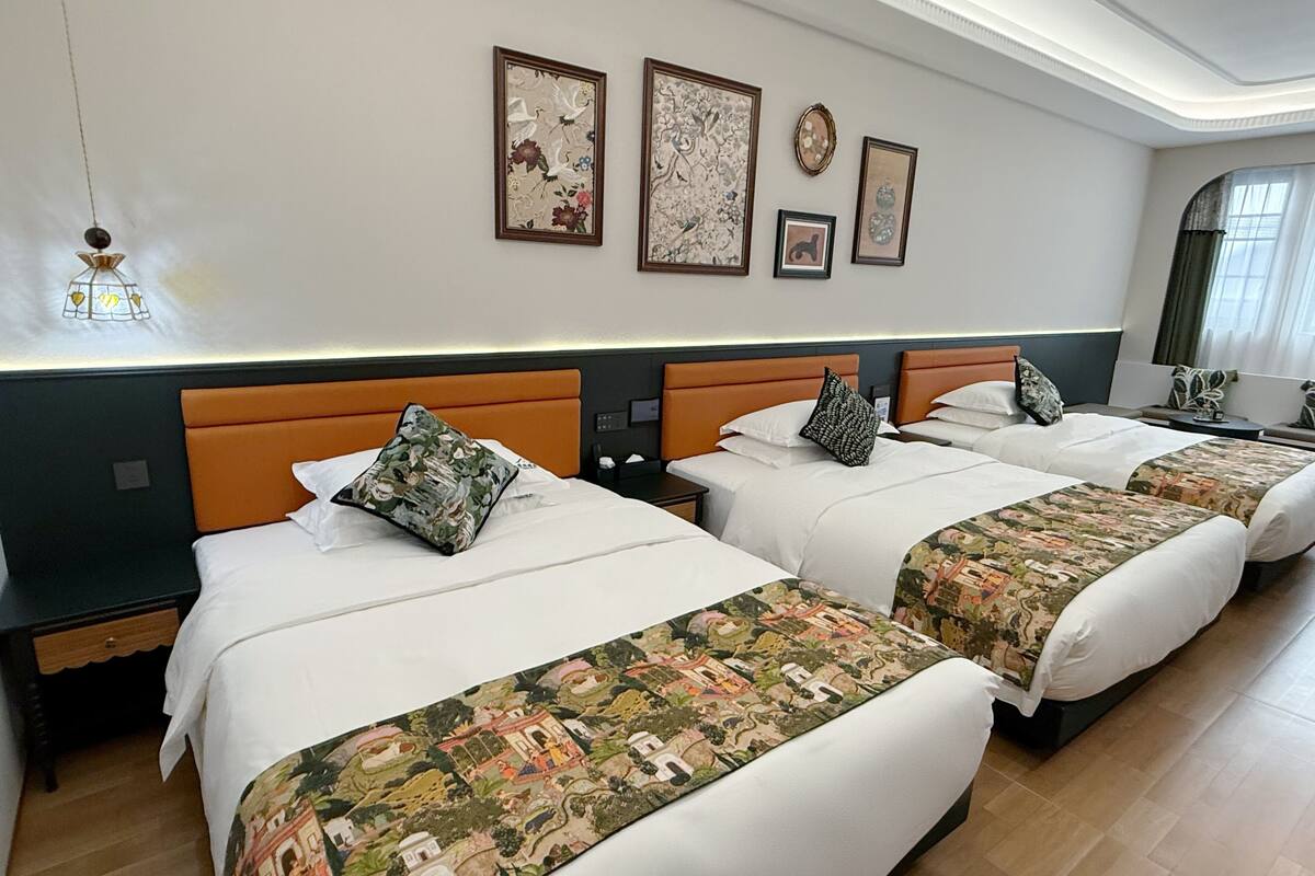 Habitación de hotel con tres camas, cada una con una colcha de estampado diferente. Hay obras de arte colgadas en la pared sobre las camas, y una pequeña mesa con una lámpara a la izquierda.