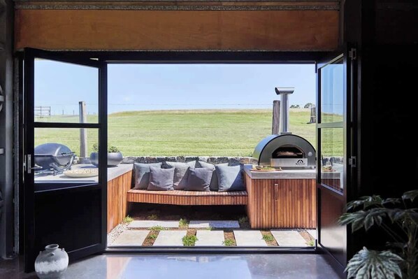 An open sliding door leading to an outdoor seating area with a grill and a pizza oven.