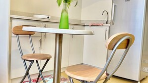 Een compacte keuken met witte kastjes, een roestvrijstalen koelkast, een kleine eettafel met een groene vaas en twee opklapbare stoelen.