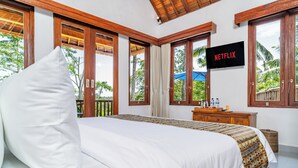 A bedroom with a large bed, wooden windows, a TV, and a view of greenery outside. - Villa Kuma Ubud (Tegallalang)