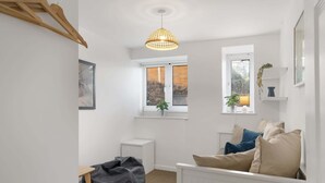 Ein kleiner, gut beleuchteter Raum mit einem weißen Sofa, einem Sitzsack, einer Pendelleuchte und zwei Fenstern mit Jalousien.