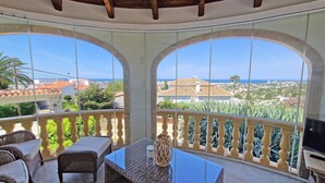 A balcony with wicker furniture and a table, overlooking a coastal town with houses and the sea. - Casa Esmick - Denia, Costa Blanca (Denia)