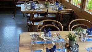Ein Restaurant mit Holztischen und -stühlen, gedeckten blauen Servietten und Tellern, und großen Fenstern mit Blick auf Grünflächen und Berge.