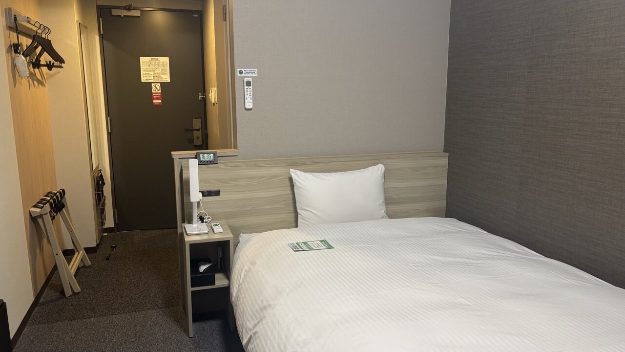 Een hotelkamer met een bed, nachtkastje, spiegel, deur en kapstok.