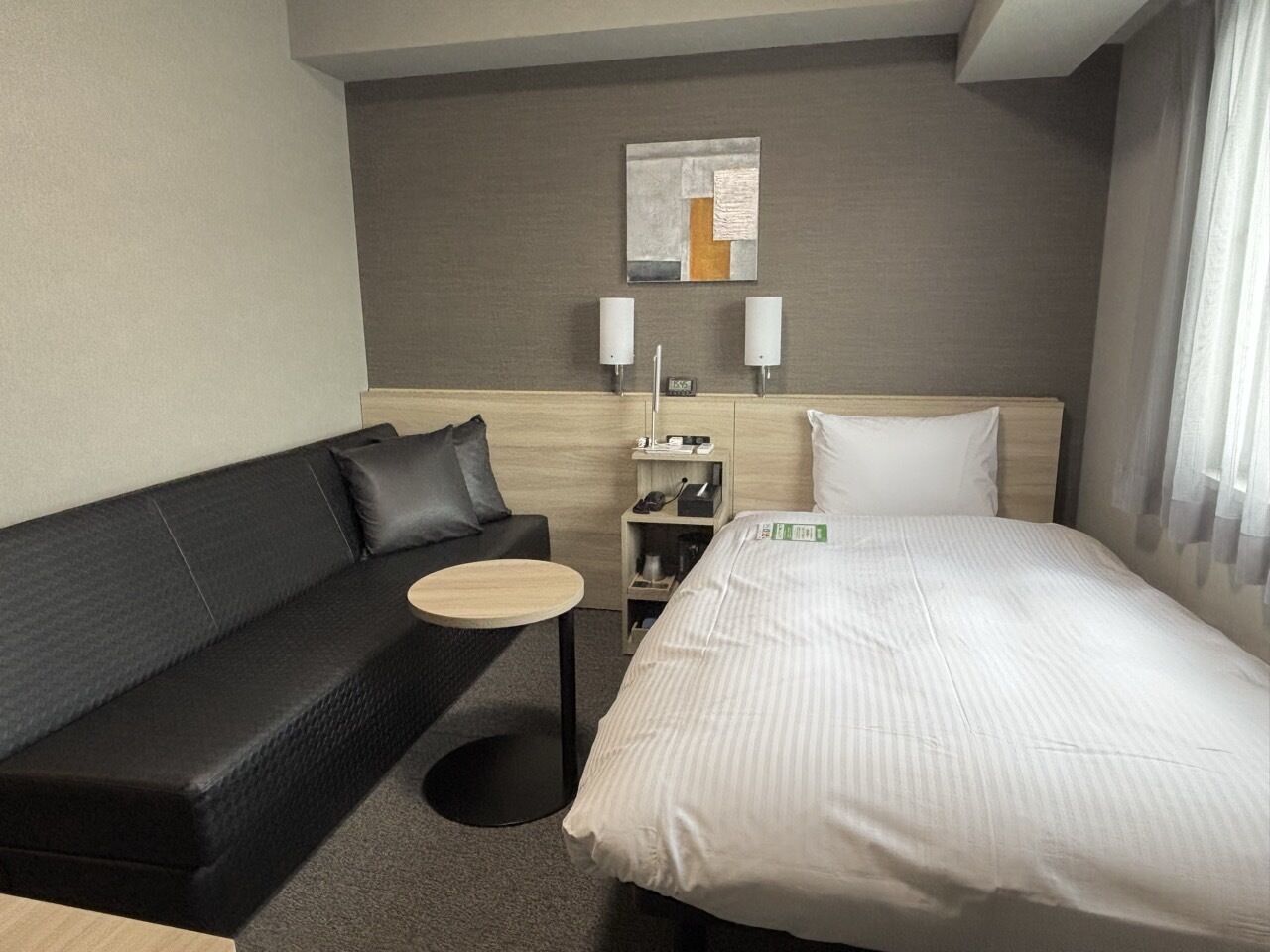 Een hotelkamer met een bed, een zwarte leren bank, een klein rond tafeltje, een nachtkastje met lamp en een kunstwerk aan de muur.
