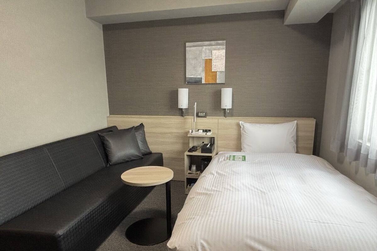 Habitación de hotel con cama, sofá de cuero negro, una mesita redonda, mesita de noche con lámpara y una obra de arte colgada en la pared.