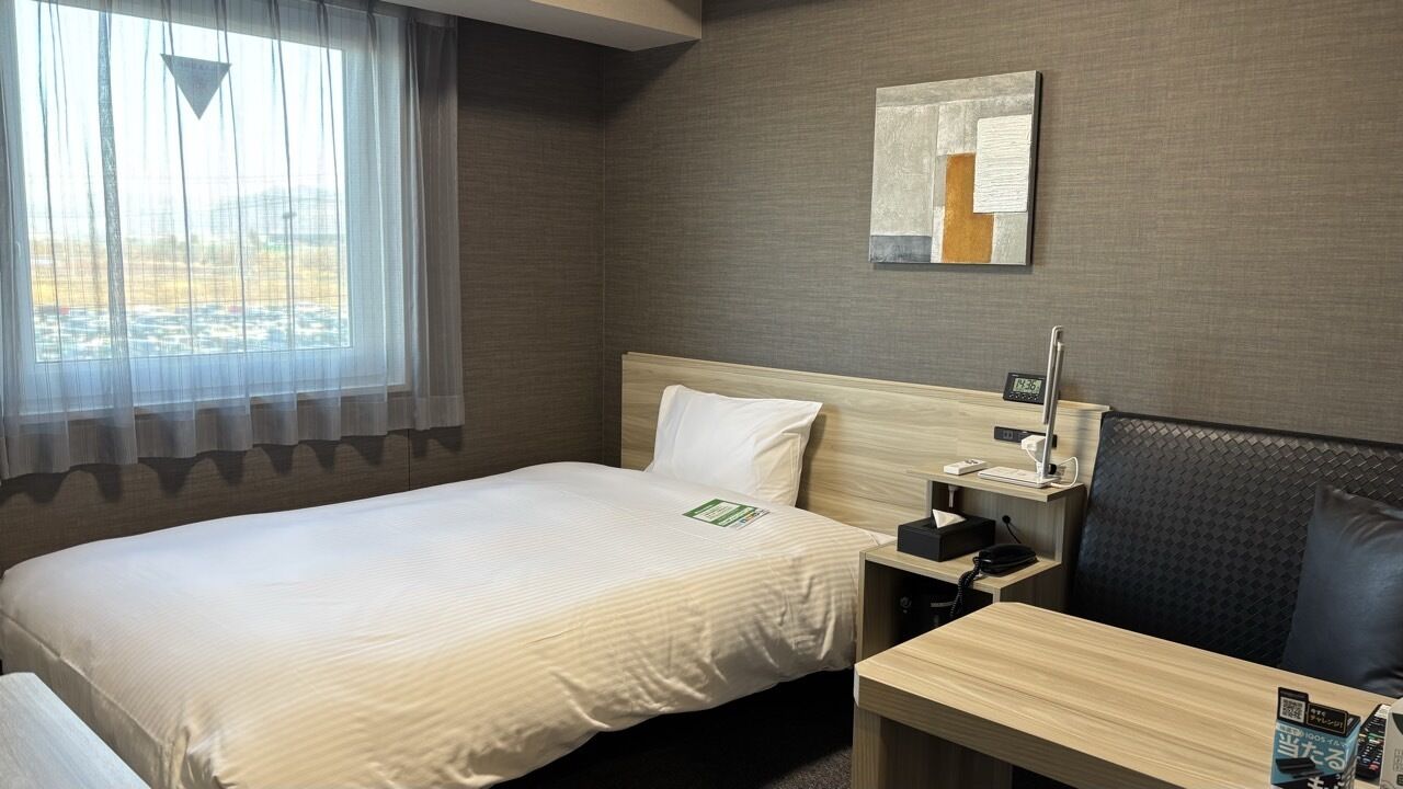Een hotelkamer met een bed, nachtkastje met telefoon, klein bureau, stoel en raam met dunne gordijnen.