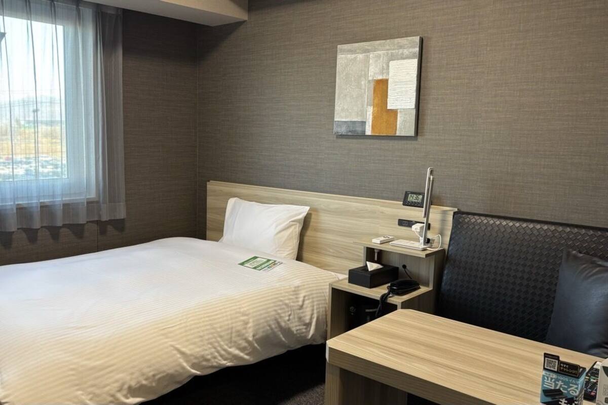 Habitación de hotel con cama, mesita de noche con teléfono, pequeño escritorio, silla y ventana con cortinas traslúcidas.