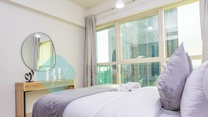 A neatly arranged bedroom with a large bed, a bedside table with a mirror, and a window with curtains. - Bloomfeilds Royal 1Br Skyscraper (Abu Dhabi)