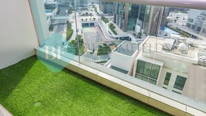 A balcony with artificial grass, a glass railing, and a view of a cityscape with modern buildings and roads. - Bloomfeilds Royal 1Br Skyscraper (Abu Dhabi)